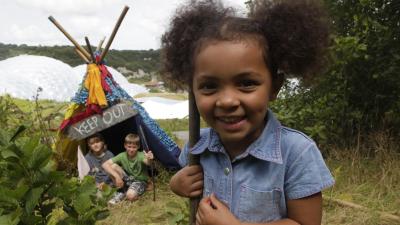 A photograph of two children hiding in a den with the Eden Project in the background and a young girl closer to the camera and holding a stick